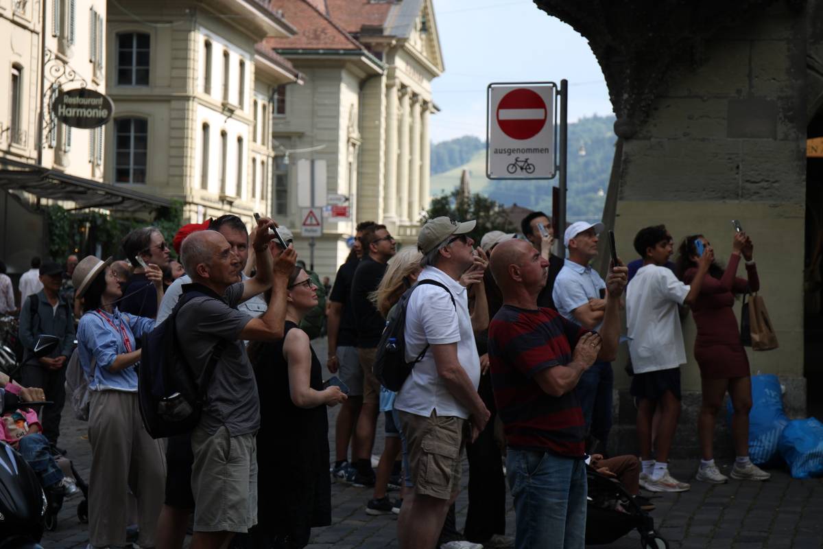 A crowd of tourists point their phones up at a church bell that is out of frame to the right
